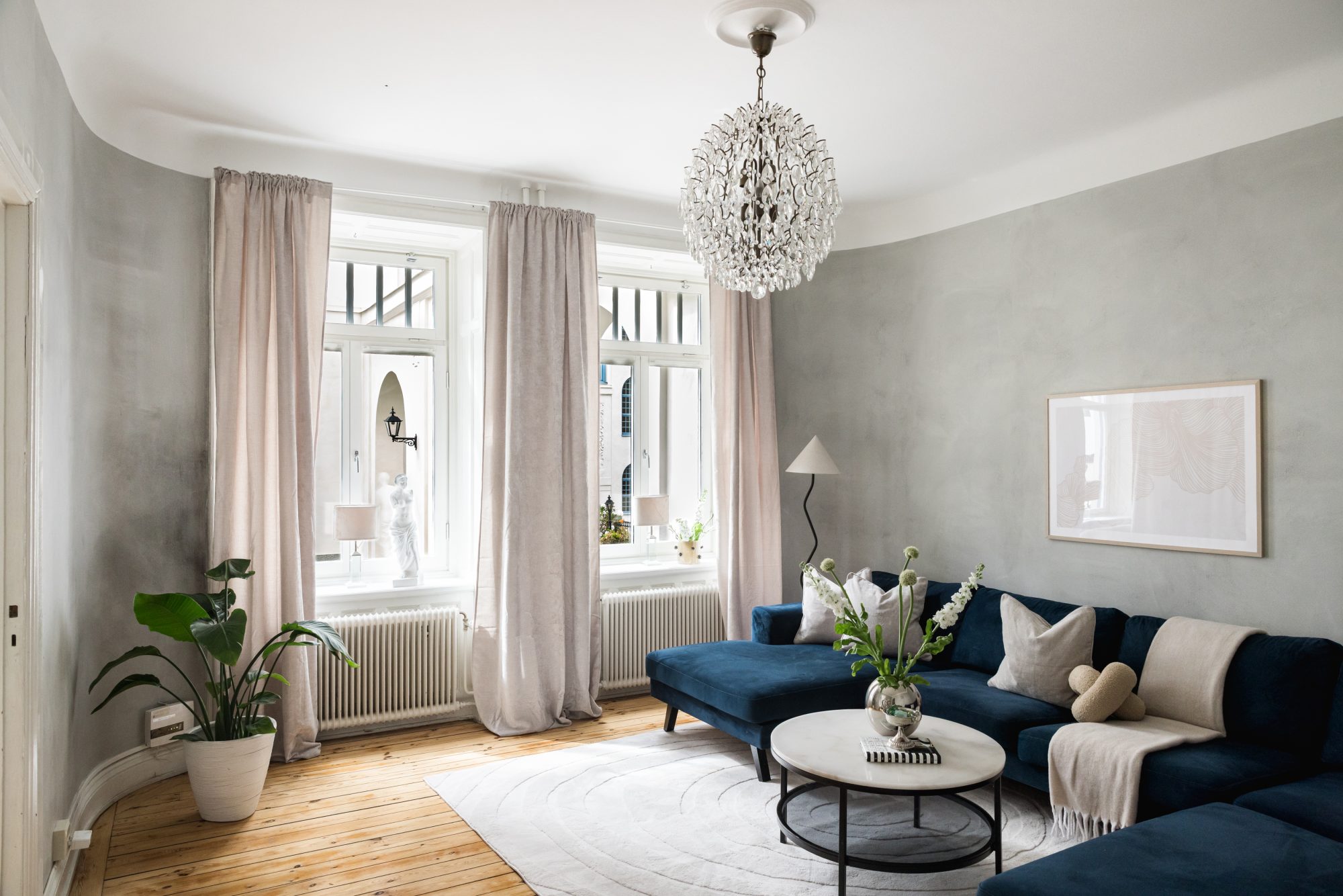 Homestylat vardagsrum på Upplandsgatan i Vasastan, Stockholm med blå sammetssoffa, kristallkrona, rosa sammetsgardiner, kalkade väggar och originalgolv i trä English: